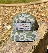 Load image into Gallery viewer, A camouflage trucker hat with a green mesh back and a white embroidered patch reading 'Folded Wing Apparel Co.' sits on a large stone outdoors with gravel and grass in the background.